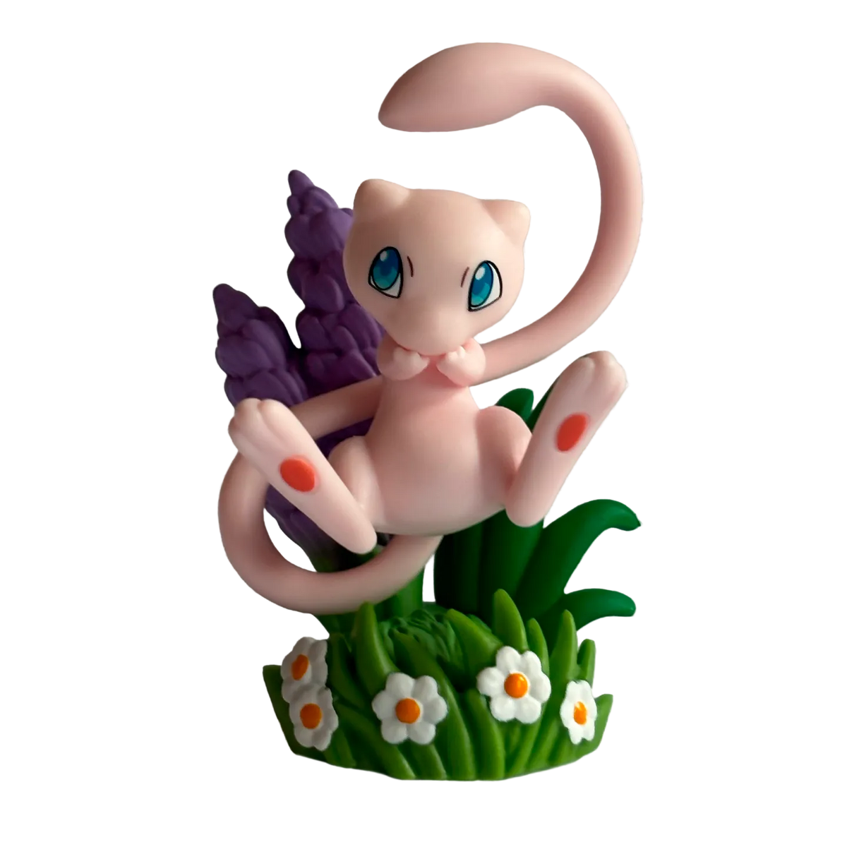 A small pink cartoon creature with large blue eyes sits among green grass, white flowers, and purple plants against a patterned background—perfect for fans of The Pokémon Company’s Pokémon TCG: 30th Celebration Figure Collection or as a display piece alongside your favorite collectibles.